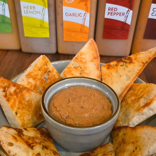 Toasted bread triangles surround a bowl of Roostr Yard's creamy red pepper dip, with sauce bottles in the background.
