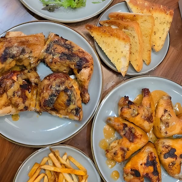 Plates of Roostr Yard grilled chicken, garlic bread, fries, and a side salad on a wooden table.